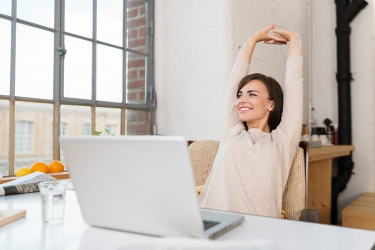 Happy relaxed young woman sitting in her kitchen with a laptop in front of her stretching her arms above her head and looking out of the window with a smile_seo-opt